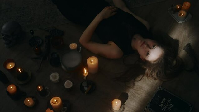 Top view of demon waking up in body of young pale lady, opening big black eyes while lying on wooden floor among burning candles during invoking ceremony indoors