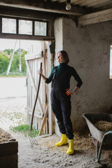 A beautiful woman farmer dressed in overalls with pitchforks in her hands is working in the cowshed.