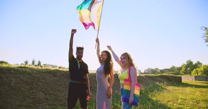 Trans woman rise and wave LGBT Rainbow Flag Standing Up for Love and Equality whit friends whit fist in air. Diversity in pride gay month. Dolly zoom out camera movement and sunset in the background.