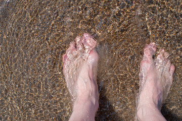 Female feet barefoot in waves of foamy water on a sandy beach on a summer day, top view of female feet, woman on the beach getting her feet wet in warm sea water, carefree vacation by the sea, 