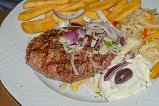Grilled bifteki - Greek meatballs with French fries, salad and tzatziki, garnished with raw onions, typical dish in Greek restaurant or fast food grill, selected focus