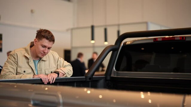 Male customer choosing pickup truck in car dealership. Young man buyer looking around big open trunk of car. Guy visitor walking in modern auto showroom and choosing new vehicle to buy or rent.
