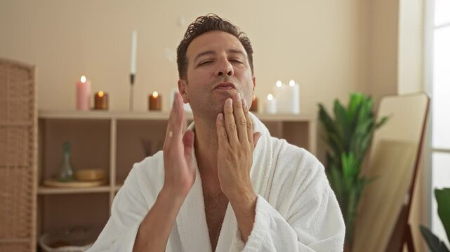 Handsome man in a white bathrobe touching his face after shaving indoors at a spa wellness center surrounded by candles and plants