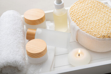 Concept of facial serum or elixir. Still life with white color jars and bottles on white wood tray, candle burning and rolled towel in bath room.