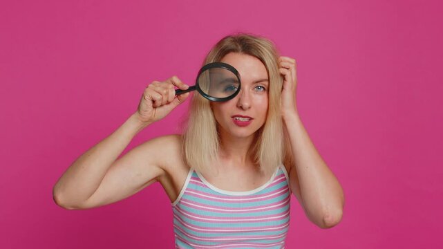 Investigator researcher scientist young woman holding magnifying glass near face, looking into camera with big zoomed funny eye, searching, analyzing. Amazed excited girl isolated on pink background.