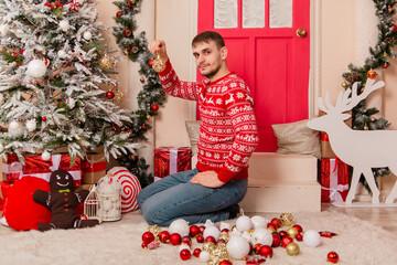 A young man in a red sweater with reindeer decorates a Christmas tree on the porch.