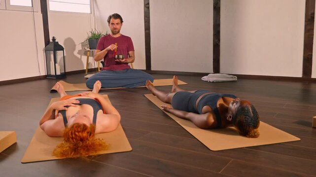 A yoga instructor using a singing bowl during a relaxation session with two participants lying on mats.