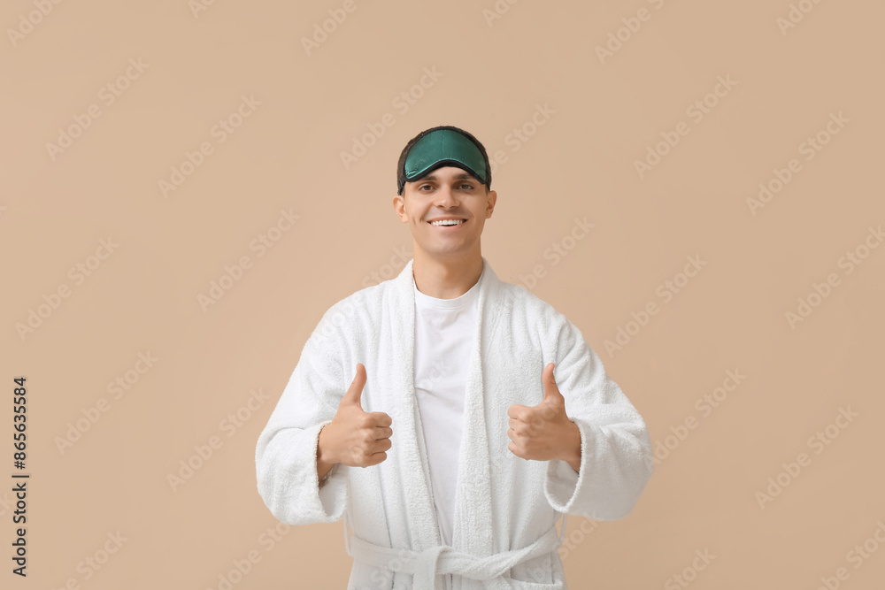 Sticker Portrait of handsome young man in bathrobe and sleeping mask showing thumbs-up on beige background