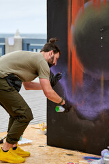 Attractive male artist is painting picture with paint spray can spraying it onto canvas at outdoor street exhibition, side view of man art maker