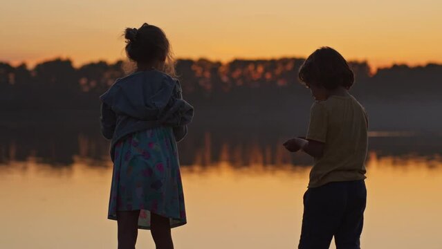 Children throwing stones into the water standing on the river bank during sunset. boy and girl standing with their backs to the camera against the backdrop of sunset, happy children having fun outside