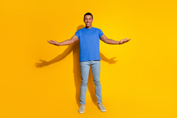 Full length photo of handsome young guy stretch hands hold empty space dressed stylish blue garment isolated on yellow color background