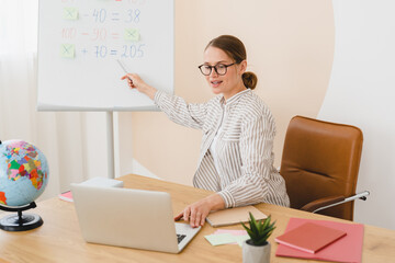 New normal. Maths distant remote online lesson. Successful young teacher tutor lector working on distance during lockdown, explaining sums on whiteboard using laptop for video call.