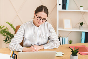 Confident young caucasian businesswoman freelancer teacher tutor headmaster working on laptop, writing projects, planning startup in office remotely.