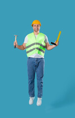 Young male builder jumping with hammer on blue background