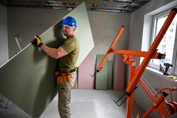 Construction worker carrying a large plasterboard with the help of a drywall lift, on a...