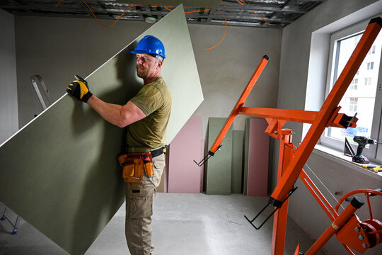 Construction worker carrying a large plasterboard with the help of a drywall lift, on a construction site