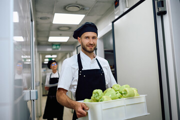 Smiling chef carrying fresh groceries from  pantry room and looking at camera.