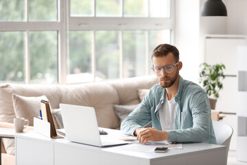 Fototapeta premium Young businessman in eyeglasses doing paper work at workplace with modern laptop in office