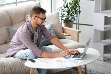 Young businessman in eyeglasses working with laptop and documents on sofa in office