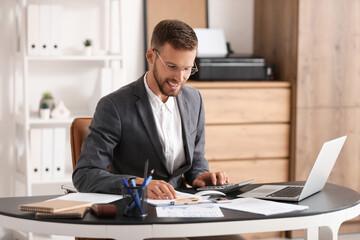 Young businessman in eyeglasses working with documents and laptop at workplace in office