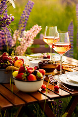Romantic table decor for a loving couple on blooming meadow with purple lupines. Two glasses of wine, flowers in a vase, silverware, fruits, wooden furniture. Sunset, golden hour, atmospheric light