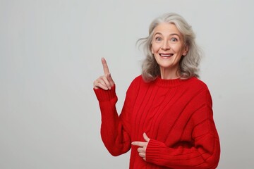 A senior woman with grey hair smiles and points to the right while wearing a red sweater against a white background.