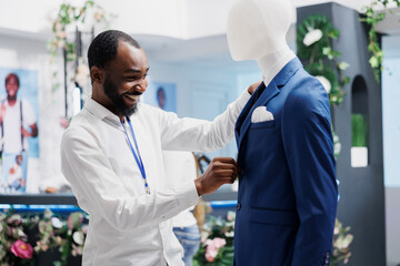 Boutique assistant fastening buttons on mannequin wearing formal jacket, displaying trendy male clothes. African american man dressing dummy model in suit in shopping mall