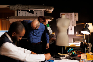 Needleworker helping senior client try on unfinished suit garment while professional fashion designer picks best adjustments to be made. Couturiers helping elderly costumer with custom made attire