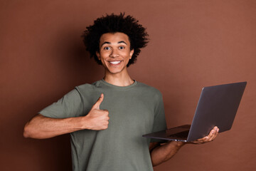 Photo of young guy in gray t shirt remote working holding laptop using for online business development thumb up isolated on brown color background