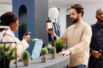 Man customer holding smartphone showing mobile discount code to saleswoman while shopping in clothing store. Cashier scanning promo coupon while serving shopper at checkout counter