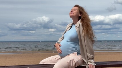 Young pregnant happy woman sitting on bench at the beach, sea and stroking her big belly