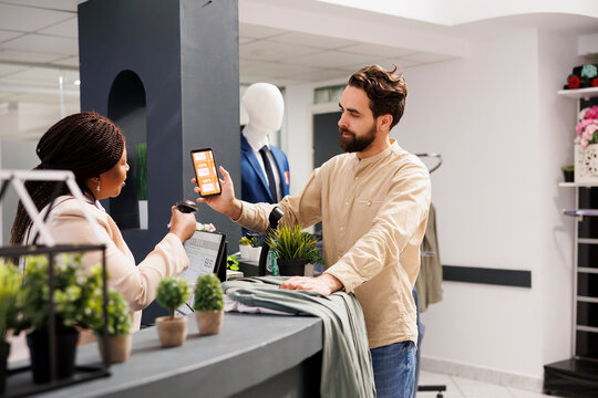African American woman cashier scanning Black Friday mobile promo code from customer smartphone, using barcode scanner. Young man shopper using coupon to get discount while shopping in clothing store