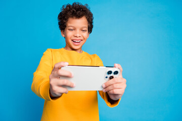 Photo portrait of small charming boy hold gadget play mobile game wear trendy yellow outfit isolated on blue color background © deagreez