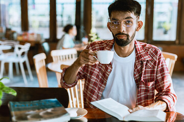 Portrait of a bearded Asian man wearing glasses. Drinking coffee and reading at a coffee shop
