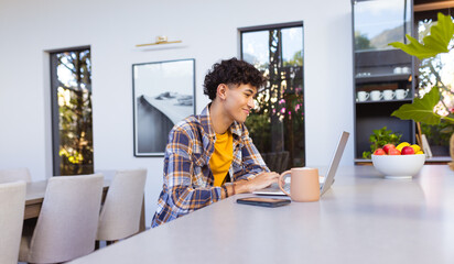 Using laptop and smiling, teenage boy working from home with coffee mug nearby