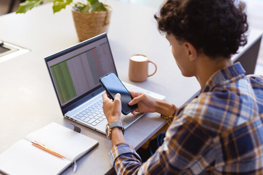Using smartphone while working on laptop, teenage boy multitasking at home office