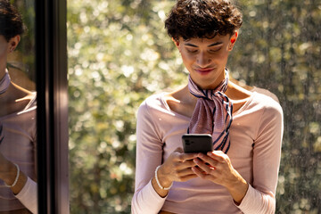 Texting on smartphone, transgender woman standing by window, smiling and relaxed