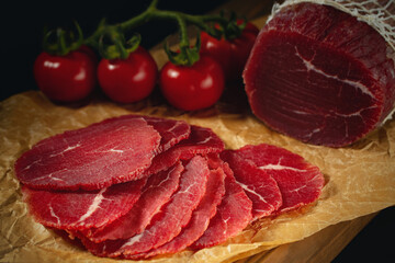 Whole and sliced bresaola on paper on a cutting board with tomatoes