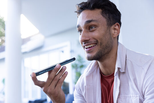 Smiling man holding smartphone and using voice assistant at home
