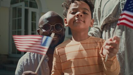 Close-up portrait of cheerful Black father and 6-year-old son, waving USA flags, posing together on front porch of family home, looking at camera and smiling, while celebrating 4th of July