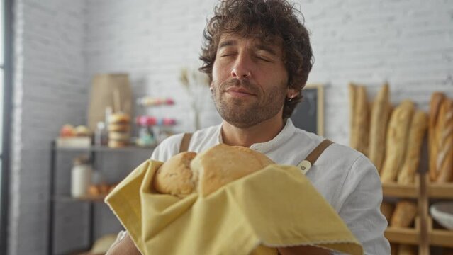 Young man holding freshly baked bread in a bakery with a content expression, surrounded by various breads and pastries in the background of a cozy, well-lit interior.