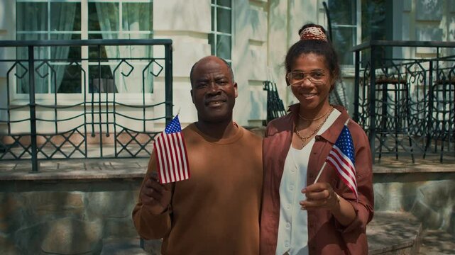 Medium full portrait of cheerful senior African American granddad embracing teenage granddaughter, waving USA flags, posing together on front porch of family home on Independence day - Powered by Adobe