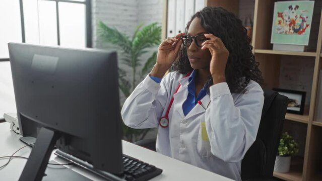 African american female doctor wearing glasses and working on a computer in a hospital clinic room with a stethoscope around her neck.