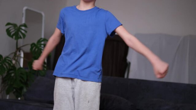 boy in a blue shirt and gray sweatpants dances at home on a wooden floor. The background features a dark sofa and a potted plant, adding a cozy, indoor atmosphere to the scene
