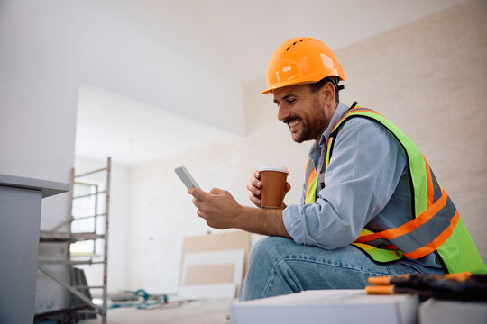 Happy worker using cell phone during coffee break at construction site. - Powered by Adobe