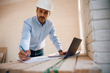 Male architect writing housing plans while using laptop at construction site.