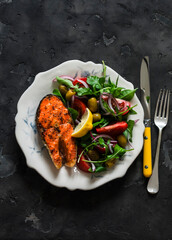 Delicious healthy lunch - grilled salmon and arugula tomatoes red onion olives salad on a dark background, top view