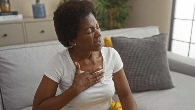 Middle-aged woman experiencing chest pain sitting indoors with a pained expression wearing a white shirt and yellow pants in a modern living room