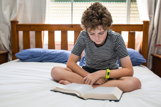 Young boy sitting cross legged on bed reading a book natural light