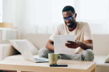 Smiling African American Man Working on Laptop in Modern Home Office Sitting on Sofa, Typing and Engaged in Cyberspace Young Millennial Freelancer Embracing Technology and Education